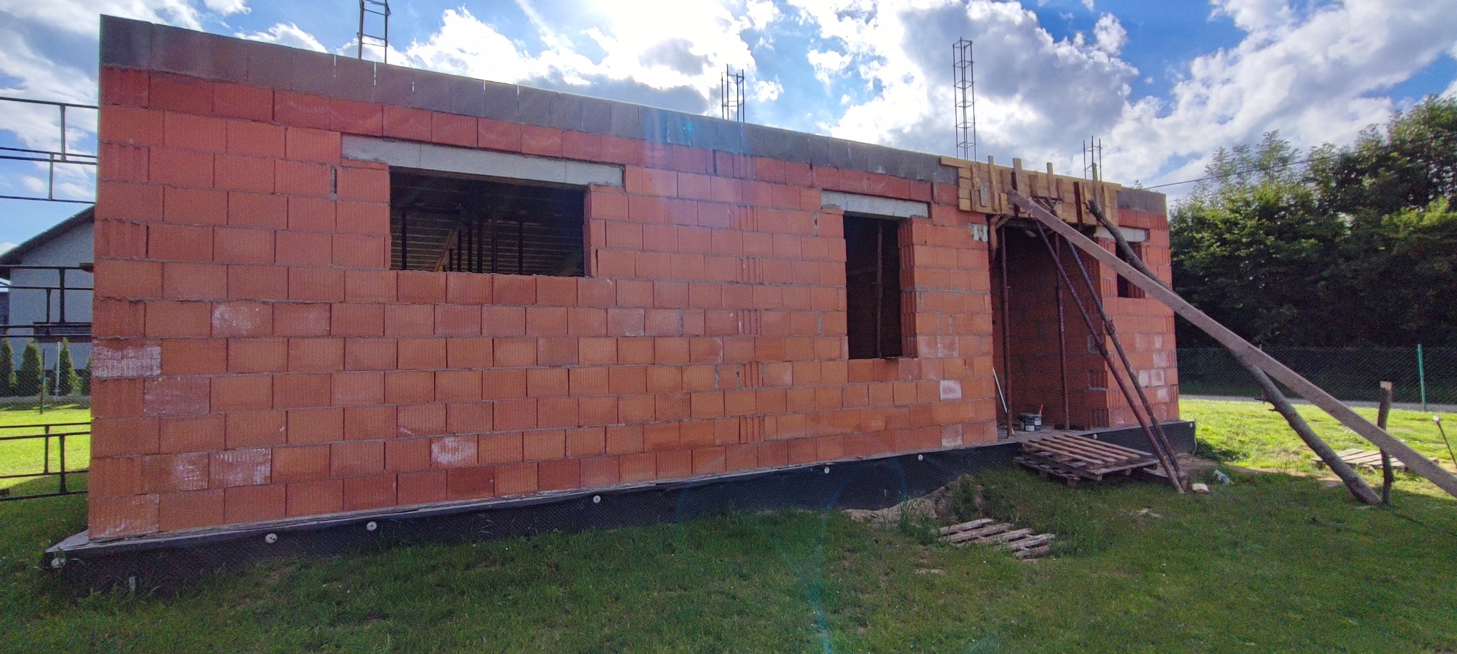 Budowa domu jednorodzinnego w trakcie wznoszenia ścian z czerwonej cegły. Widoczne otwory okienne i drzwiowe, szalunki, zbrojenia oraz rusztowania.