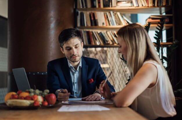 Mężczyzna w garniturze i kobieta omawiają dokumenty przy stole z laptopem i misą owoców, w tle regał z książkami.