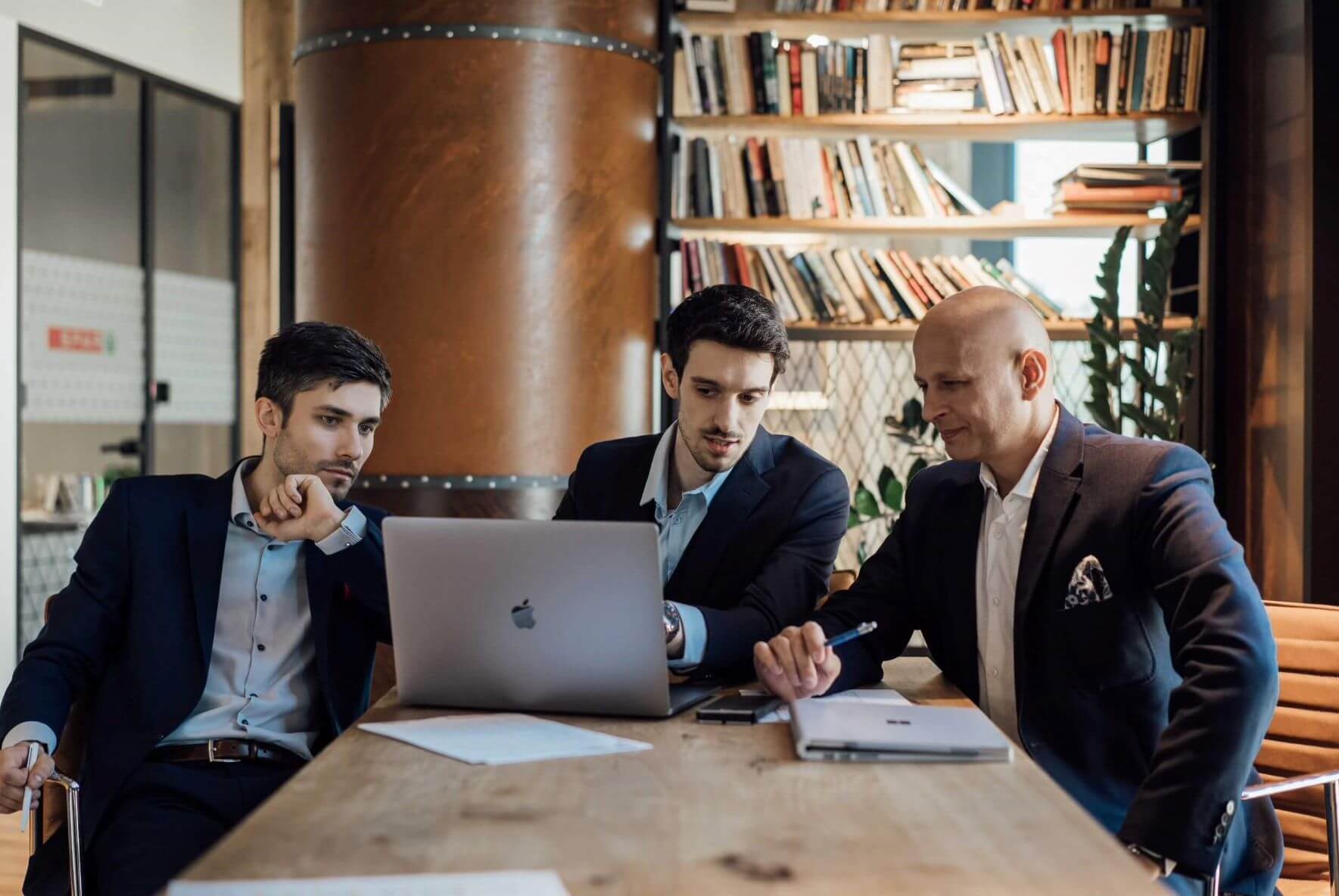 Trzech mężczyzn w garniturach analizuje dane na laptopie przy drewnianym stole w biurze, widoczny regał z książkami i dekoracyjna kolumna.