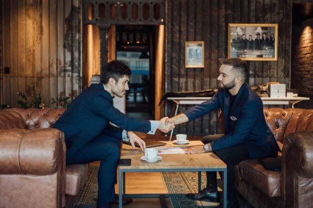 Dwóch mężczyzn w garniturach w uścisku dłoni nad drewnianym stołem z dokumentami i filiżankami kawy w eleganckim wnętrzu z skórzanymi sofami i dekoracyjnymi panelami ściennymi.
