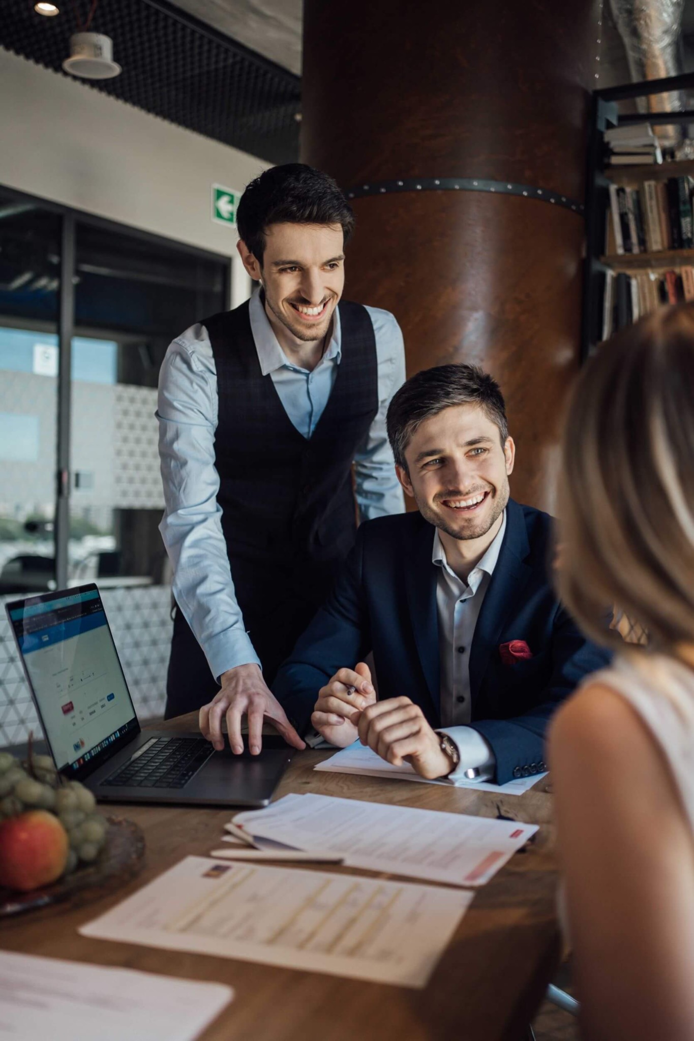 Dwóch uśmiechniętych mężczyzn w eleganckich strojach omawia dokumenty z klientką przy drewnianym stole z laptopem i owocami w tle nowoczesnego biura.