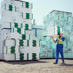 Pracownik w kasku i kamizelce odblaskowej przenosi płytę ociepleniową na tle stosów spakowanego materiału izolacyjnego na placu budowy.
