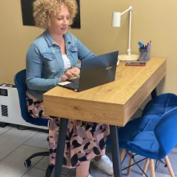 Kobieta z kręconymi włosami w dżinsowej kurtce pracuje na laptopie ASUS przy biurku z lampką, w tle grzejnik i ciemna ściana.