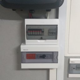 Elcom vision security - Szare urządzenie Fronius Symo nad dwiema białymi skrzynkami z bezpiecznikami i przełącznikami zamontowanymi na szarej ścianie.