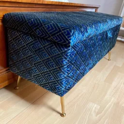 Niebieska, tapicerowana skrzynia z geometrycznym wzorem i złotymi nóżkami stoi na drewnianej podłodze obok drewnianej komody.