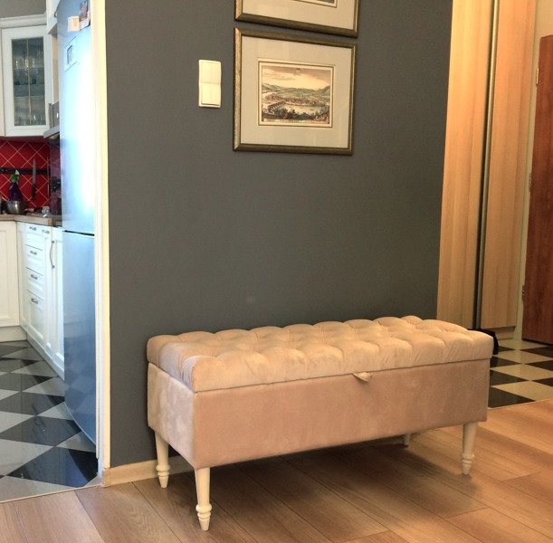 Beżowa, pikowana pufa ze schowkiem na drewnianych nóżkach, ustawiona w przedpokoju na drewnianej podłodze, z widokiem na kuchnię z białą zabudową i czarno-białą podłogą.