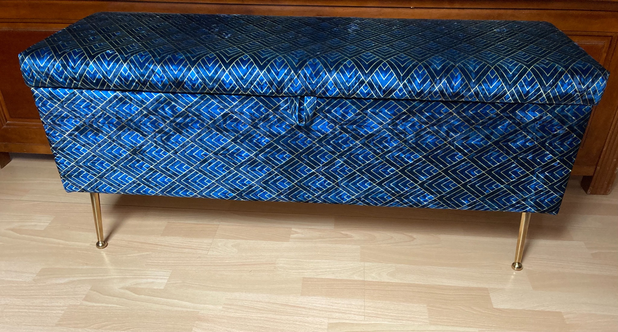 Niebieska skrzynia tapicerowana welurem ze złotym geometrycznym wzorem, na złotych nóżkach, stojąca na podłodze z paneli.