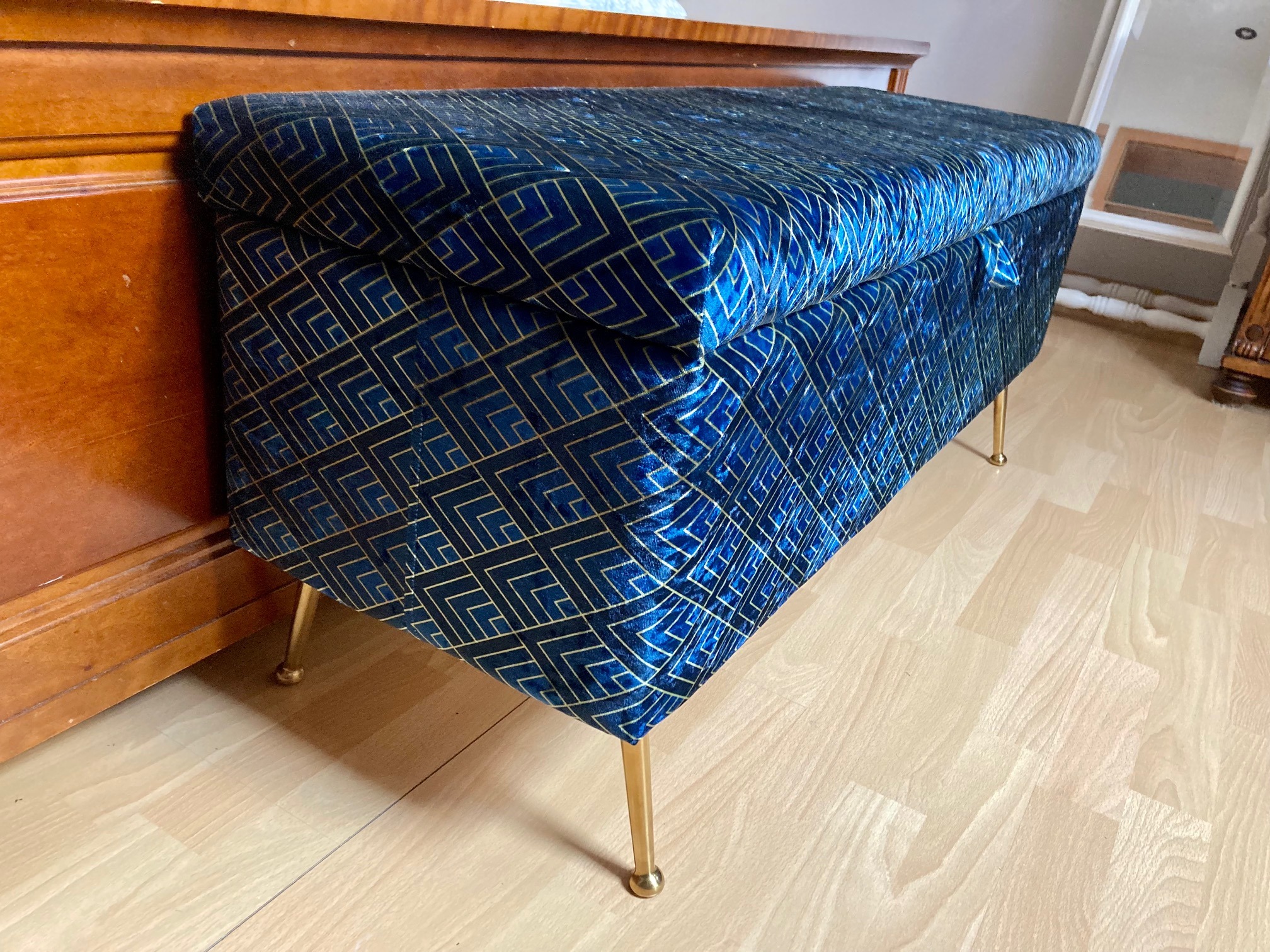 Niebieska, tapicerowana skrzynia z geometrycznym wzorem i złotymi nóżkami stoi na drewnianej podłodze obok drewnianej komody.