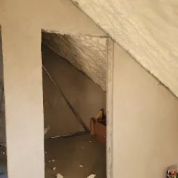 Poddasze ocieplone pianką PUR, widok na otw&oacute;r w ścianie, kartonowe pudło w głębi, paczka soku Capri-Sun przy podłodze.