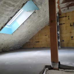 Poddasze z oknem dachowym, ocieplone szarą pianką poliuretanową, widoczna surowa ściana z bloczków i drewniany słup podpierający konstrukcję dachu.