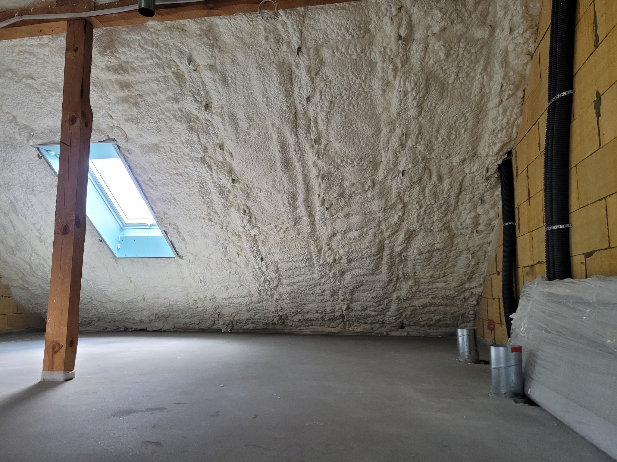 Poddasze po ociepleniu pianką: widoczna warstwa izolacji natryskowej na skosach dachu, drewniany słup podtrzymujący konstrukcję oraz okno dachowe wpuszczające światło.