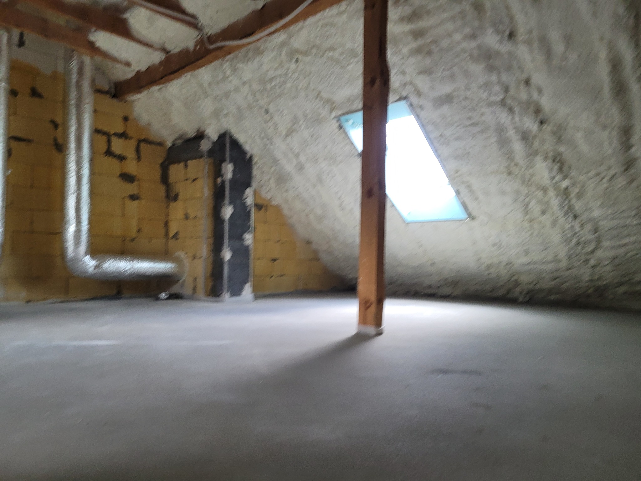 Poddasze po ociepleniu pianką PUR, widoczne krokwie, okno dachowe i fragment instalacji wentylacyjnej na ścianie, surowy beton na podłodze.