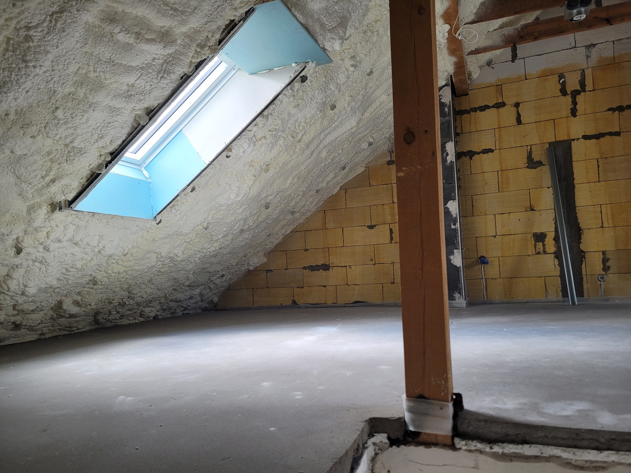 Poddasze z oknem dachowym, ocieplone szarą pianką poliuretanową, widoczna surowa ściana z bloczków i drewniany słup podpierający konstrukcję dachu.