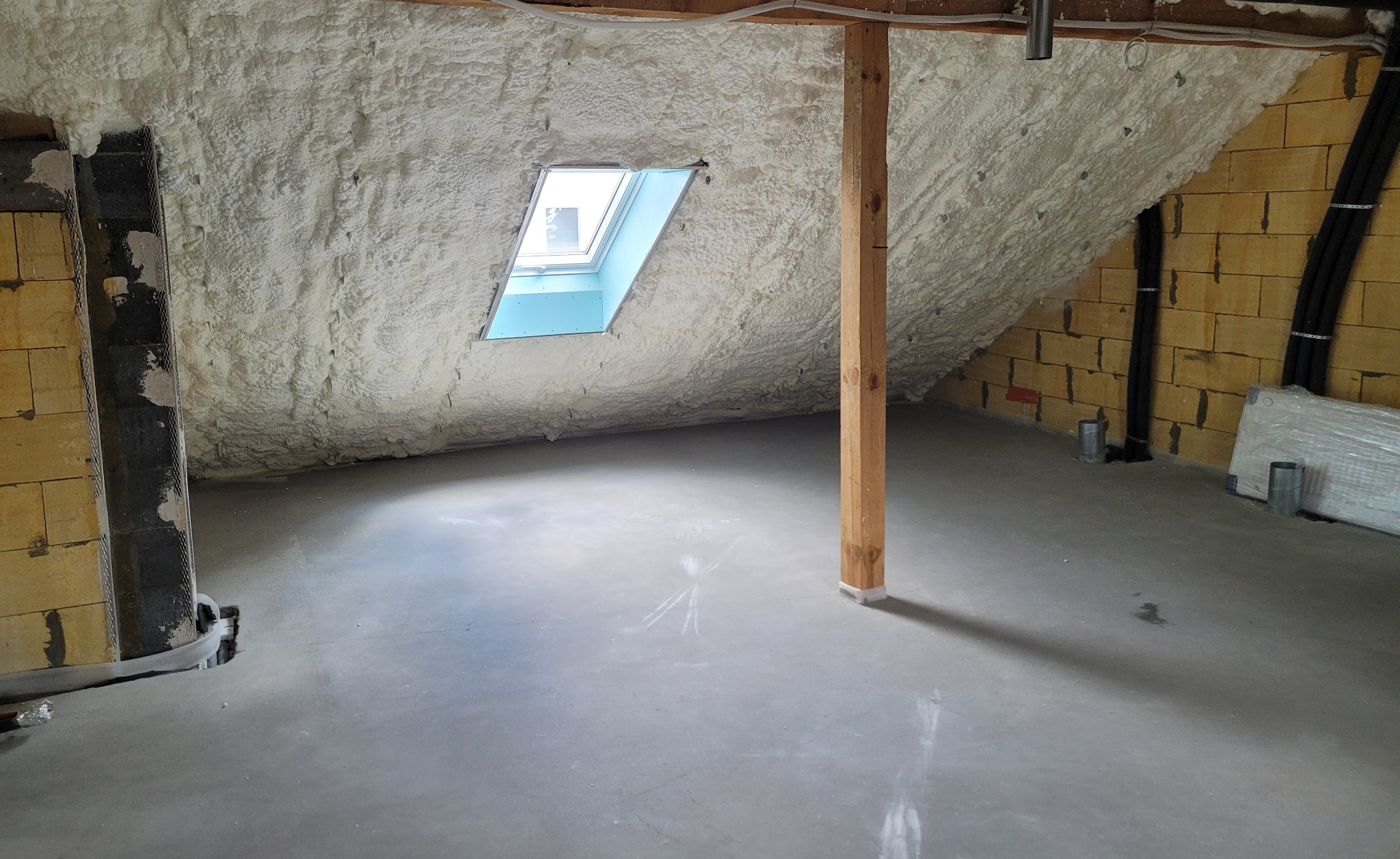 Poddasze po ociepleniu pianką PUR, widoczne okno dachowe, drewniany słup podtrzymujący konstrukcję, oraz fragment ściany z pustaków i instalacji.