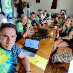 Grupa uśmiechniętych osób w koszulkach z logo firmy siedzi przy drewnianym stole, na którym leżą laptopy z widocznymi wykresami i prezentacjami dotyczącymi symboliki logo. Jedna osoba robi selfie.