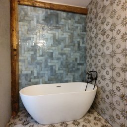 Kamiński Usługi Budowlane - Nowoczesna, wolnostojąca wanna w łazience z niebiesko-szarą mozaiką ułożoną w jodełkę i płytkami podłogowymi w kwiatowy wzór, podkreślona drewnianą belką.