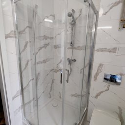 Kamiński Usługi Budowlane - Narożna kabina prysznicowa z chromowanymi profilami i drzwiami ze szkła hartowanego, zamontowana w łazience wykończonej białymi płytkami imitującymi marmur z szarymi żyłami, widoczny również...