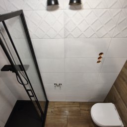 Kamiński Usługi Budowlane - Nowoczesna łazienka z kabiną prysznicową z czarnymi ramami, białe płytki z geometrycznym wzorem na ścianach, imitacja drewna na podłodze i ścianie, widoczne otwory na gniazdka elektryczne.
