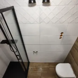 Nowoczesna łazienka z kabiną prysznicową z czarnymi ramami, białe płytki z geometrycznym wzorem na ścianach, imitacja drewna na podłodze i ścianie, widoczne otwory na gniazdka elektryczne.