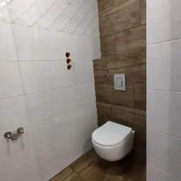 Nowoczesna łazienka z białymi i drewnopodobnymi płytkami, wisząca toaleta z przyciskiem spłukiwania, widoczne otwory na armaturę.