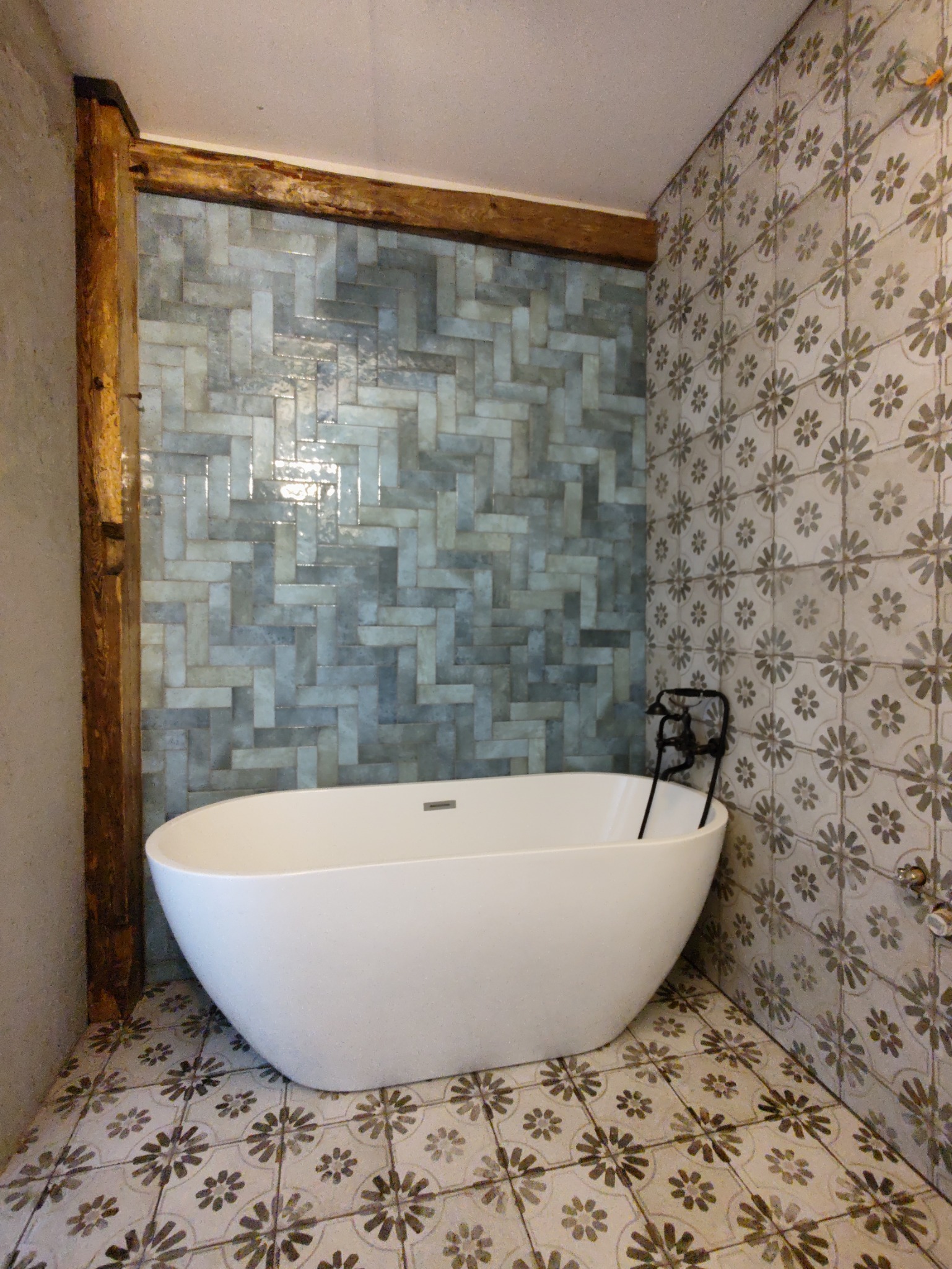 Nowoczesna, wolnostojąca wanna w łazience z niebiesko-szarą mozaiką ułożoną w jodełkę i płytkami podłogowymi w kwiatowy wzór, podkreślona drewnianą belką.