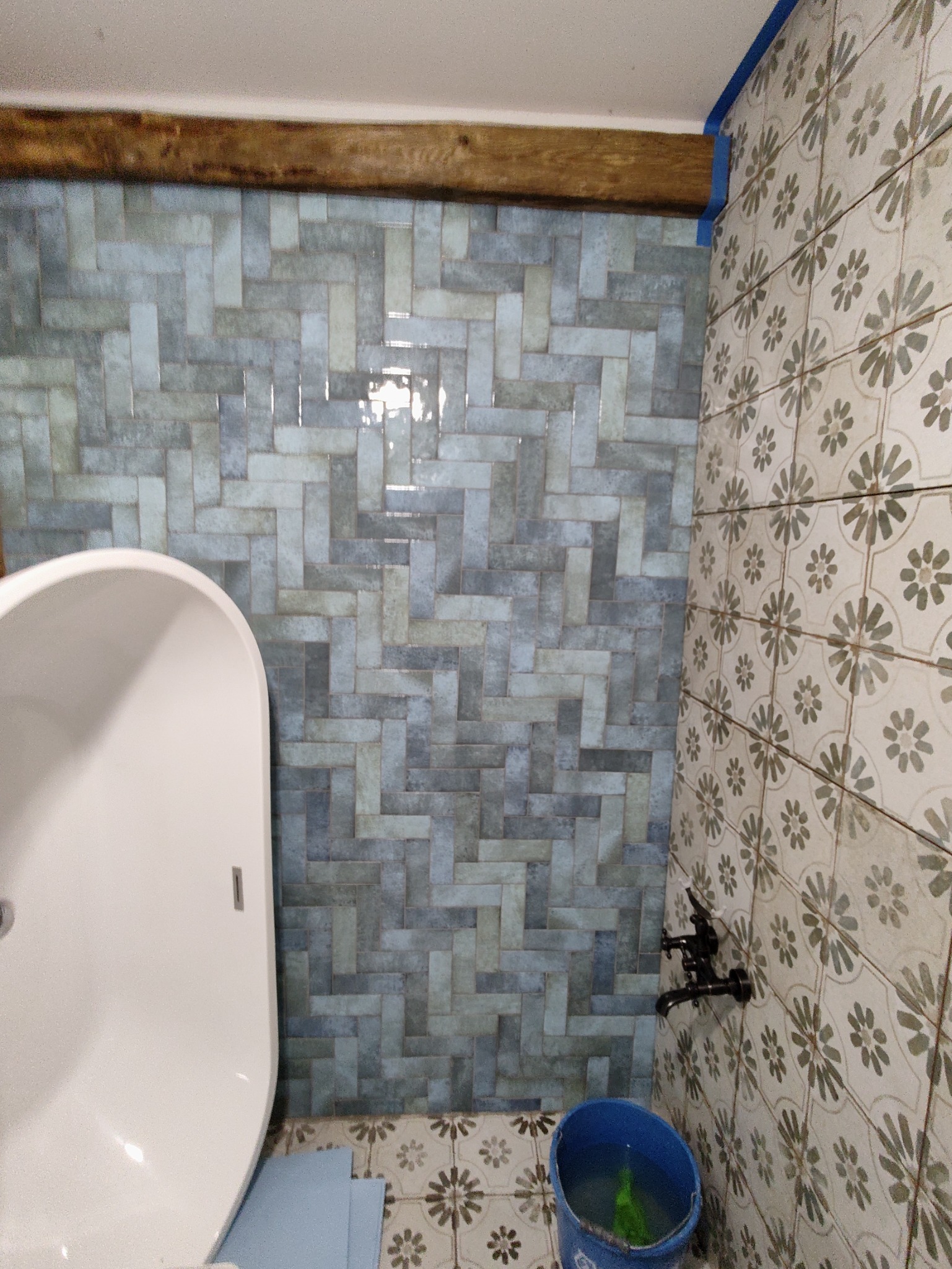 Fragment łazienki z wanną, niebieskimi płytkami ułożonymi w jodełkę na ścianie, wzorzystymi płytkami na podłodze i ścianie, oraz drewnianą belką pod sufitem.