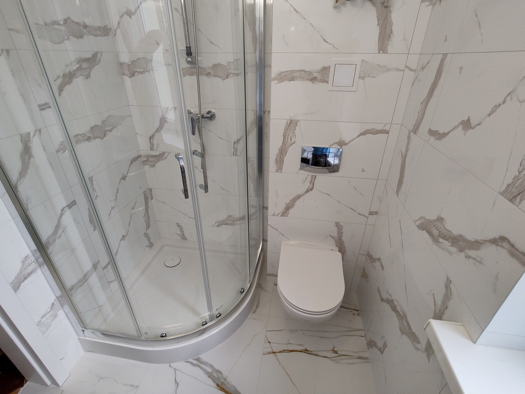 Nowoczesna łazienka z kabiną prysznicową z chromowanymi detalami i podwieszaną toaletą, wykończona białymi płytkami z marmurkowym wzorem.