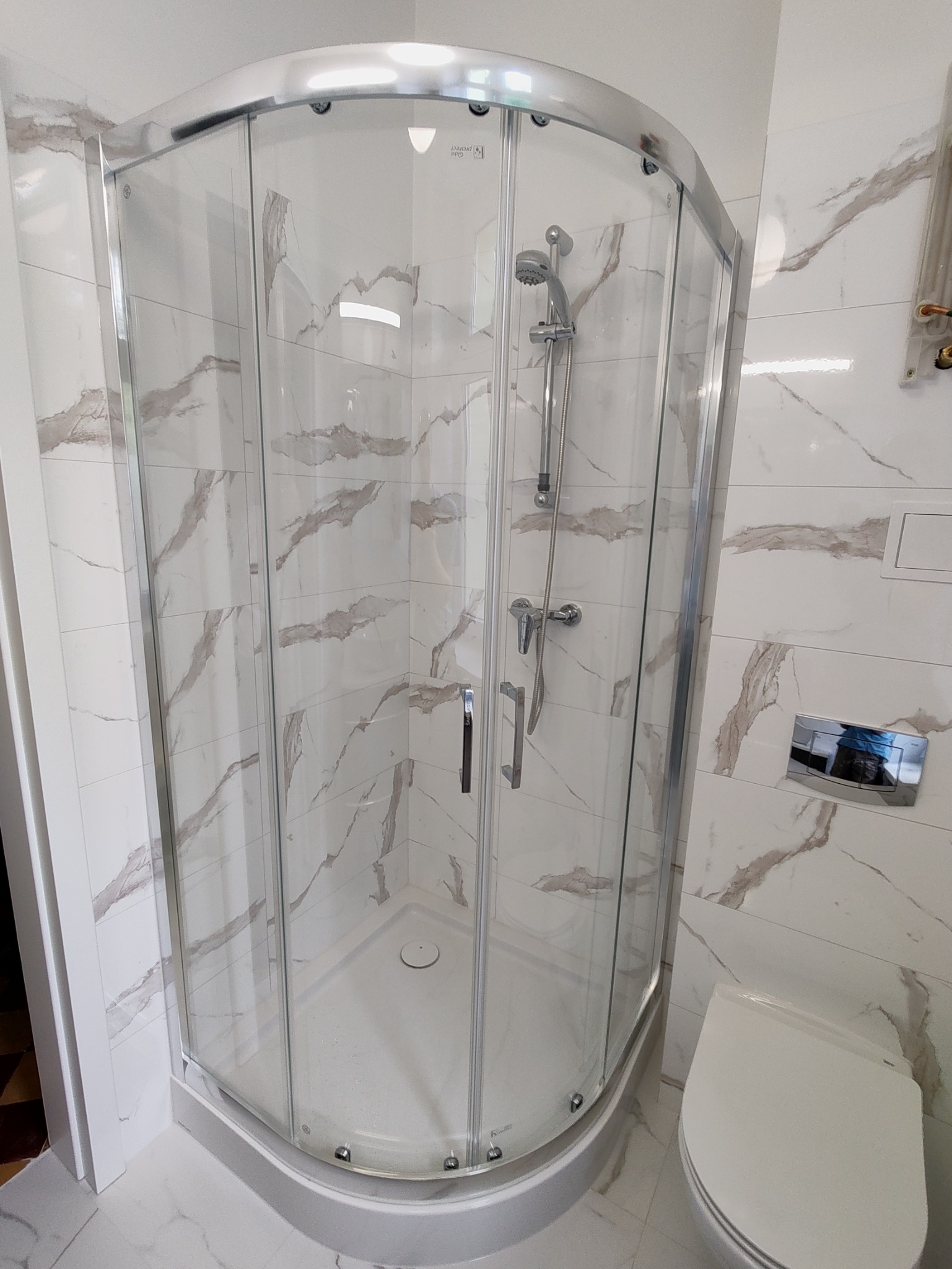 Narożna kabina prysznicowa z chromowanymi profilami i drzwiami ze szkła hartowanego, zamontowana w łazience wykończonej białymi płytkami imitującymi marmur z szarymi żyłami, widoczny również...