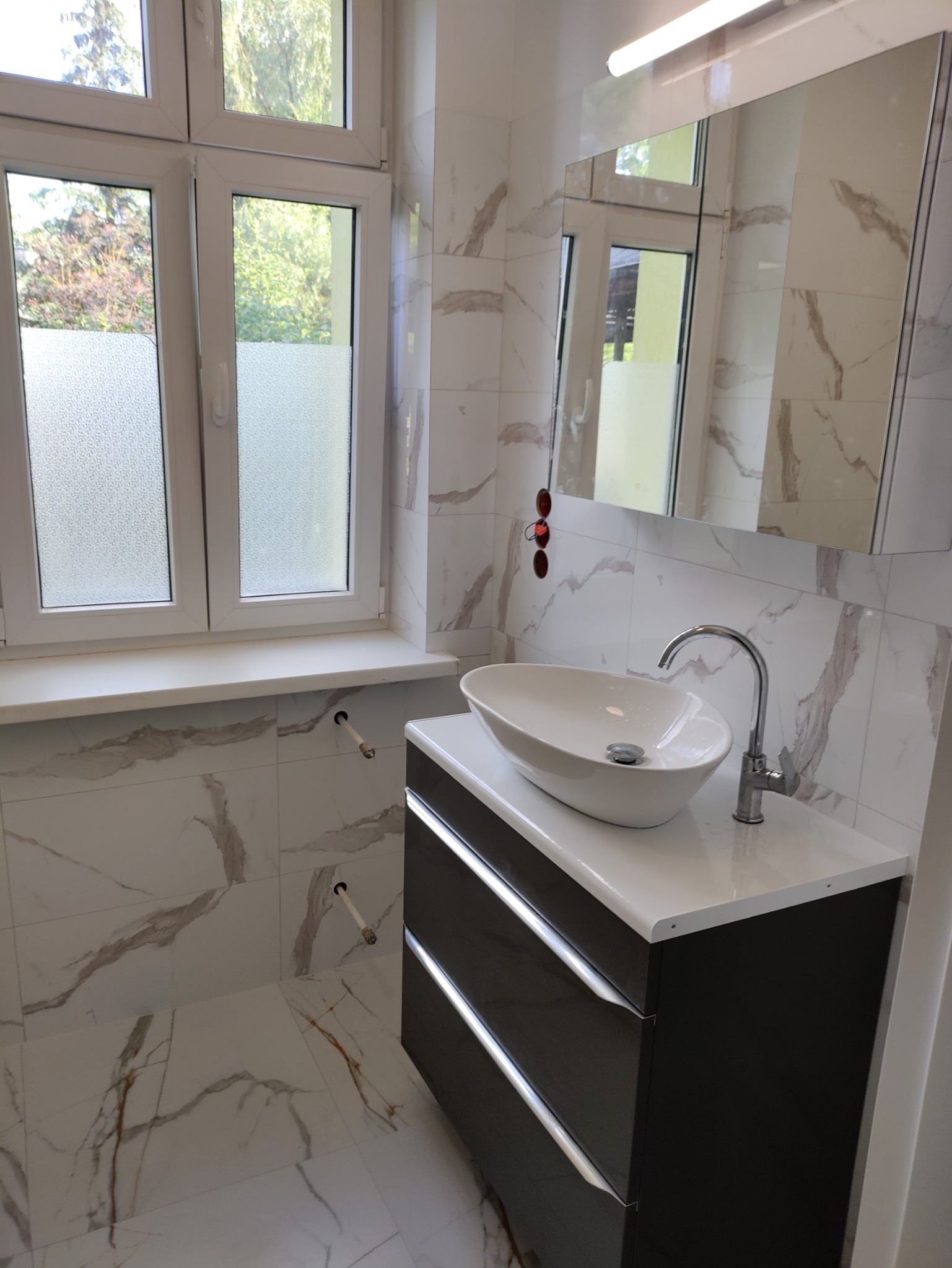 Nowoczesna łazienka w trakcie wykańczania: owalna umywalka nablatowa, szafka z ciemnymi frontami, marmurowe płytki na ścianach i podłodze, widoczne rury wystające ze ściany.