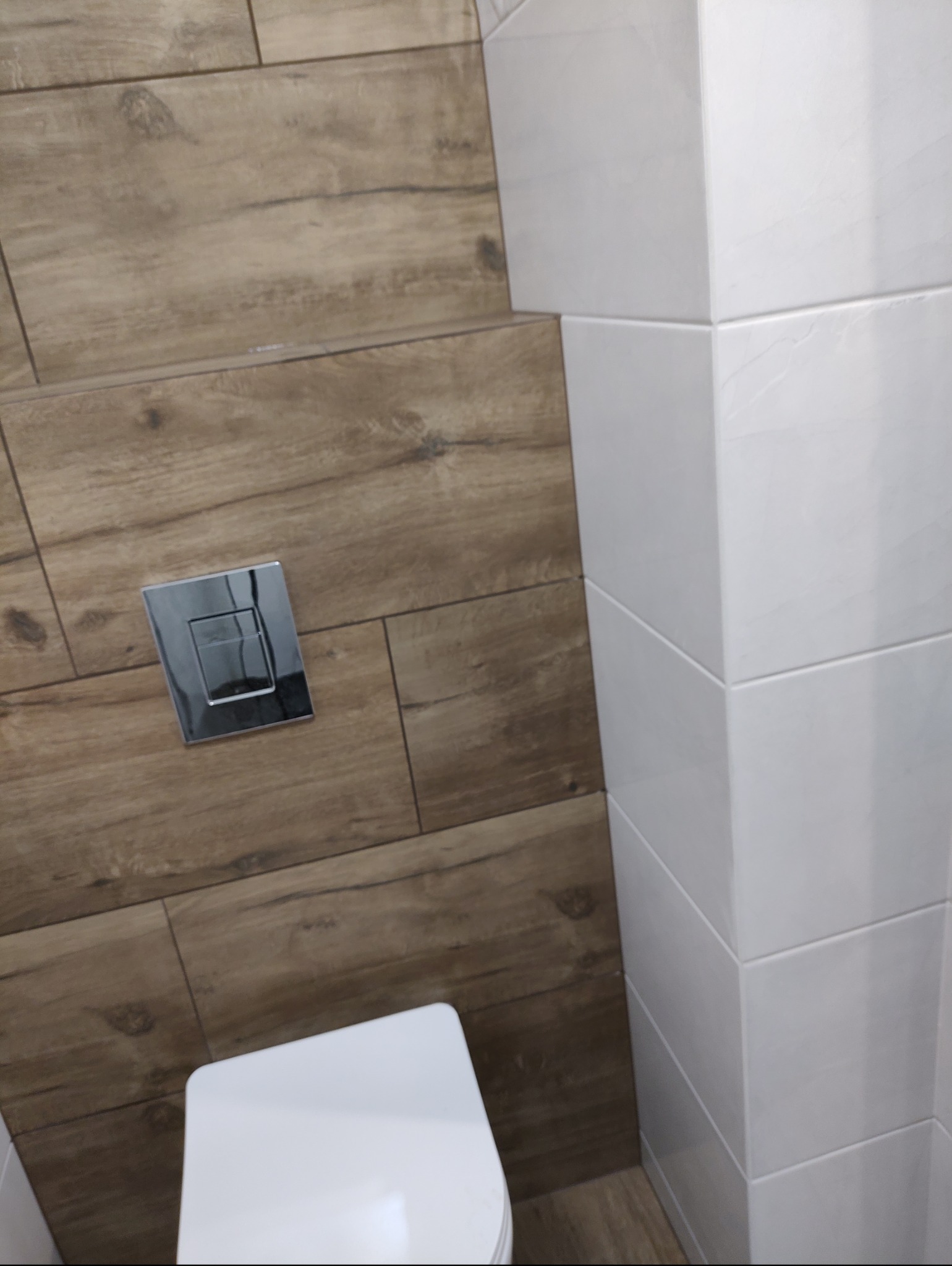 Fragment wykończonej łazienki z brązowymi płytkami imitującymi drewno, białą zabudową i chromowanym przyciskiem spłuczki.