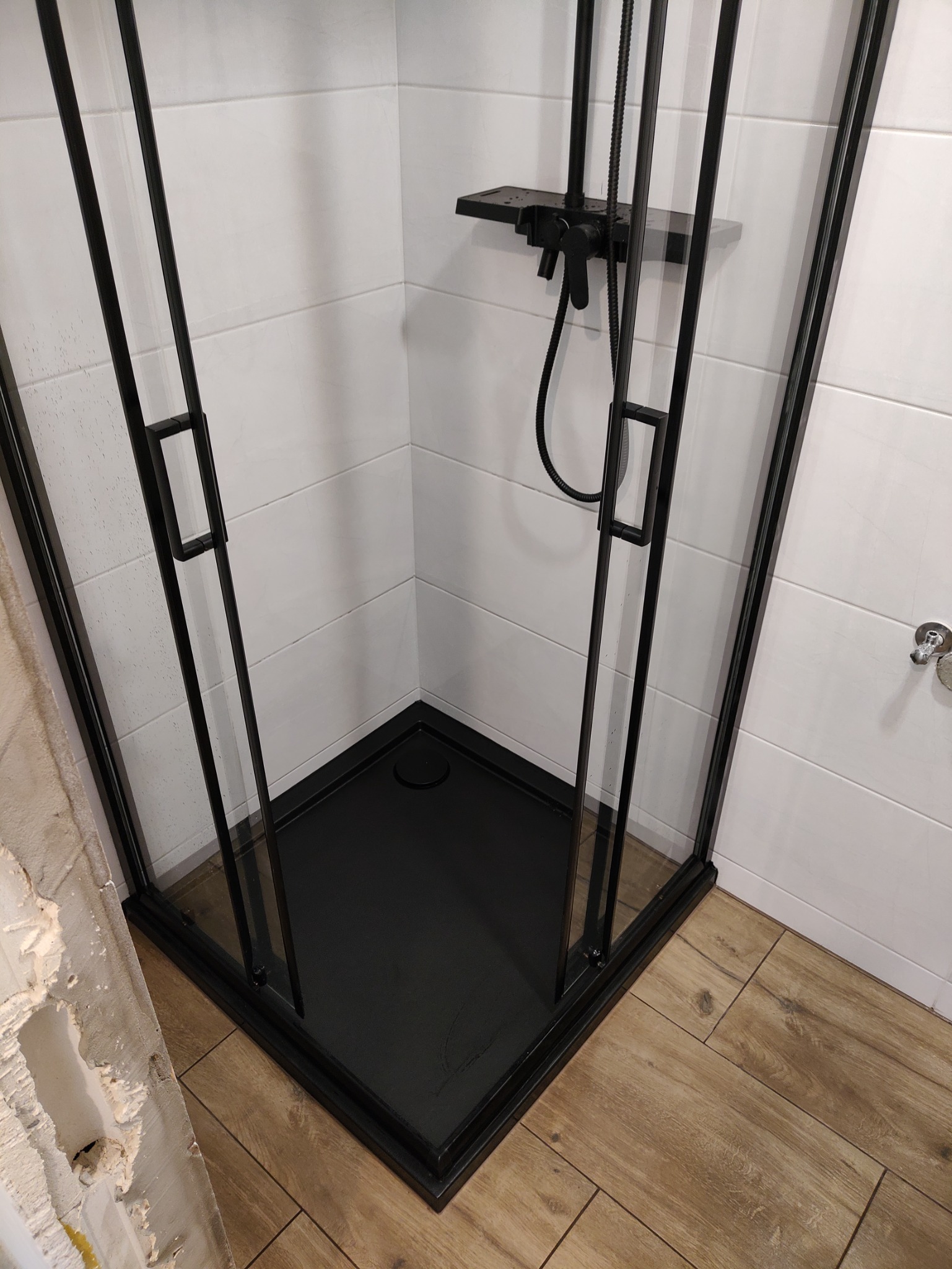 Czarna kabina prysznicowa z brodzikiem, białe płytki na ścianach, drewnopodobne panele podłogowe, widoczny fragment uszkodzonej ściany przed wykończeniem.
