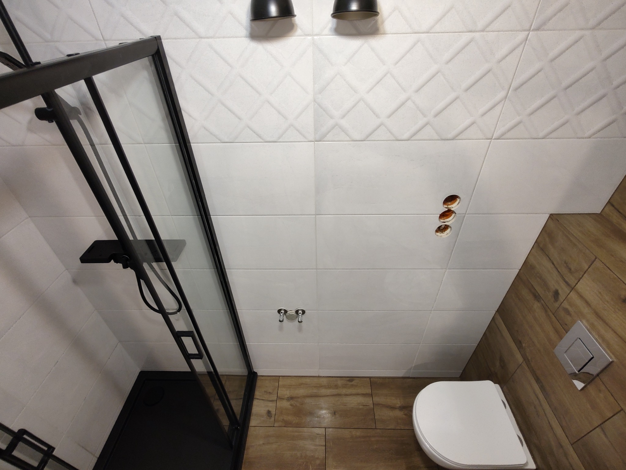 Nowoczesna łazienka z kabiną prysznicową z czarnymi ramami, białe płytki z geometrycznym wzorem na ścianach, imitacja drewna na podłodze i ścianie, widoczne otwory na gniazdka elektryczne.