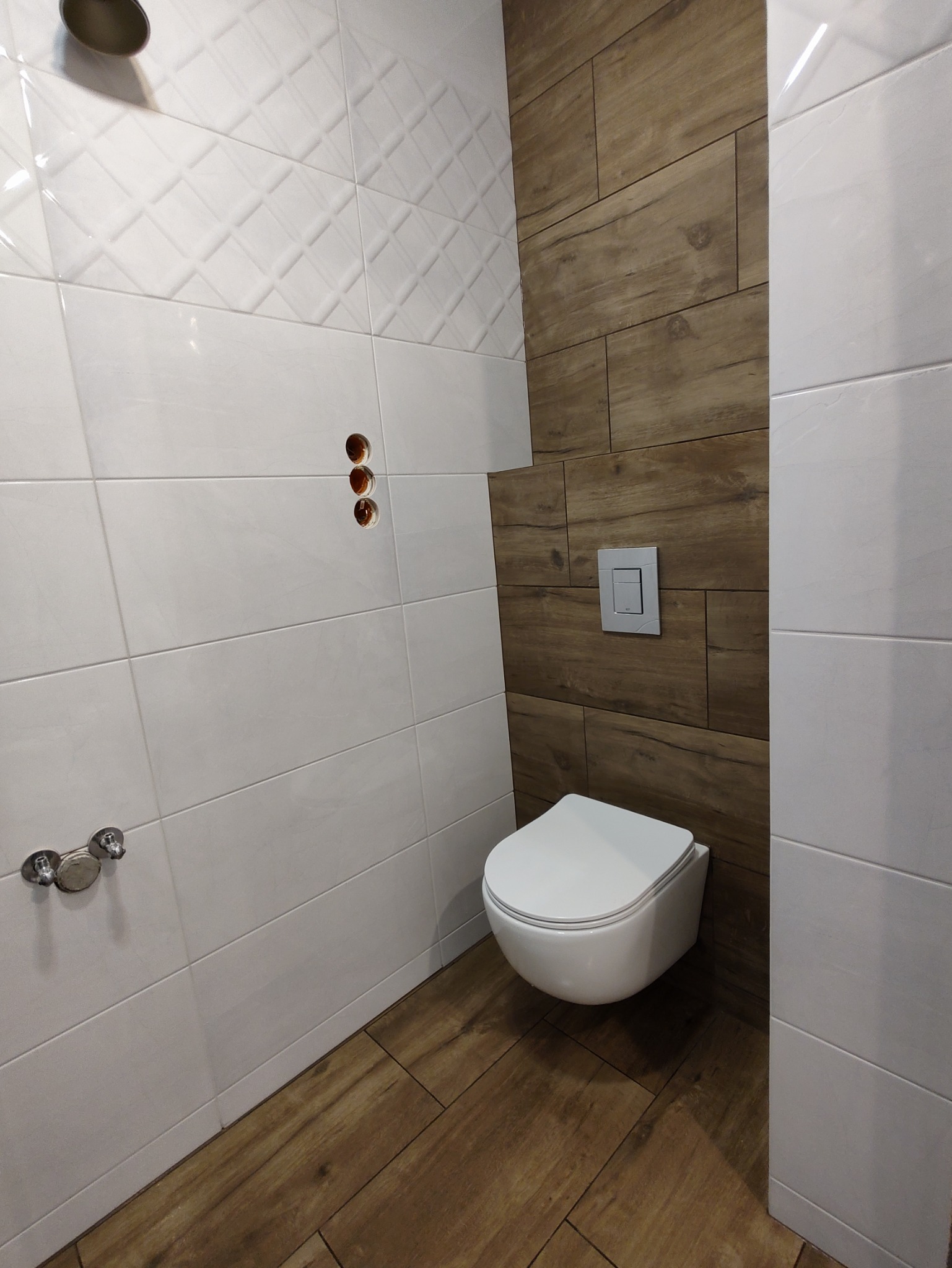 Nowoczesna łazienka z białymi i drewnopodobnymi płytkami, wisząca toaleta z przyciskiem spłukiwania, widoczne otwory na armaturę.