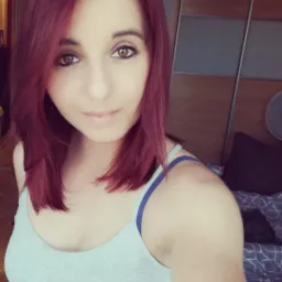 Portret kobiety z burgundowymi włosami, ubrana w szary top na tle szafy i ł&oacute;żka.