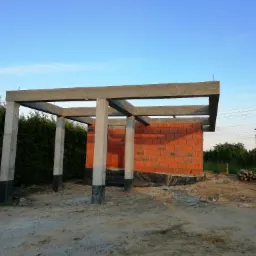 Wznoszenie ścian z czerwonej cegły na fundamencie z izolacją, widoczne betonowe słupy i elementy konstrukcyjne pod zadaszenie, teren budowy w trakcie prac.