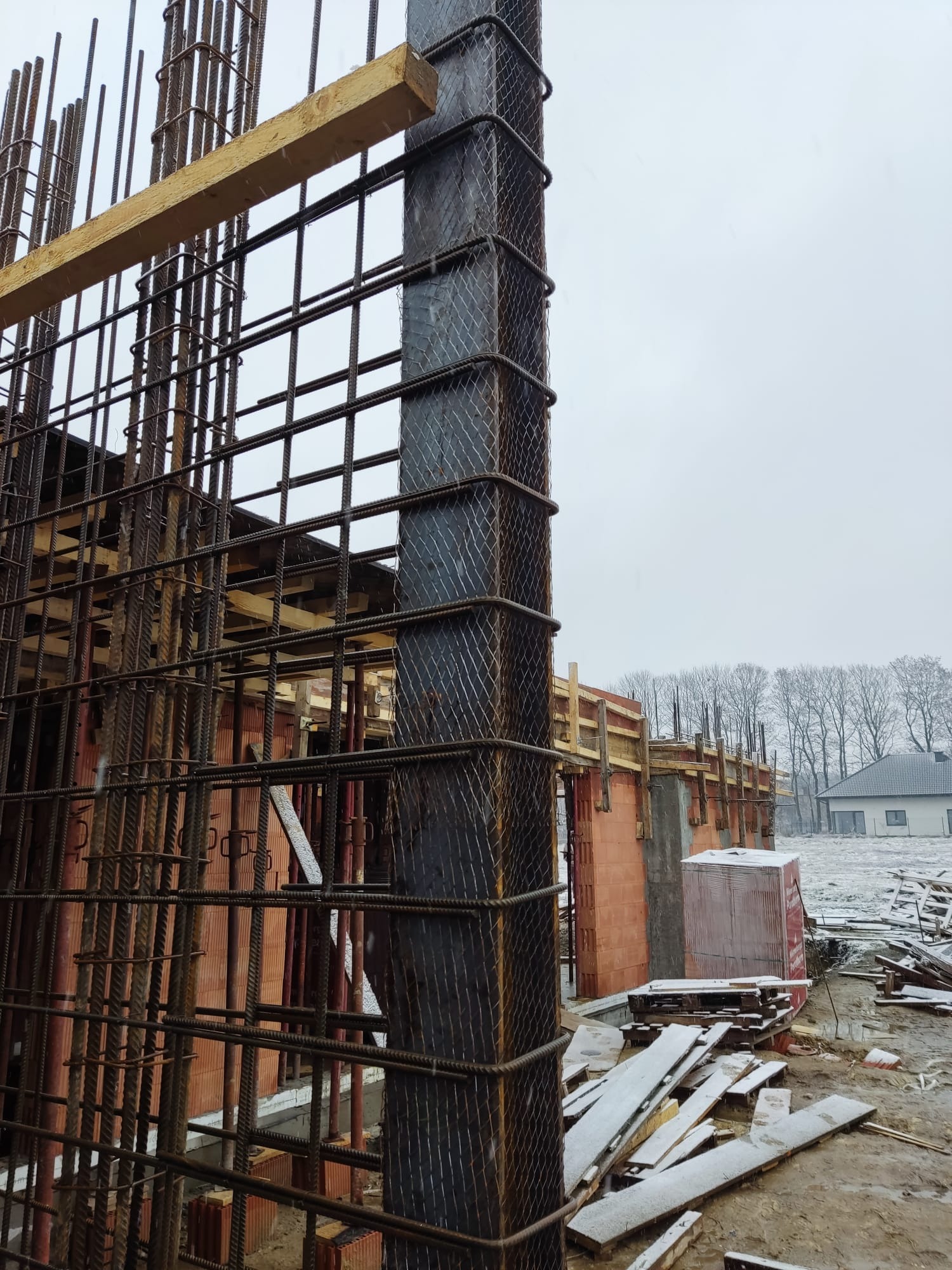 Wiązanie zbrojenia pionowych słupów na budowie domu z czerwonej cegły, widoczne drewniane szalunki i przygotowane deski na ziemi, lekki śnieg.
