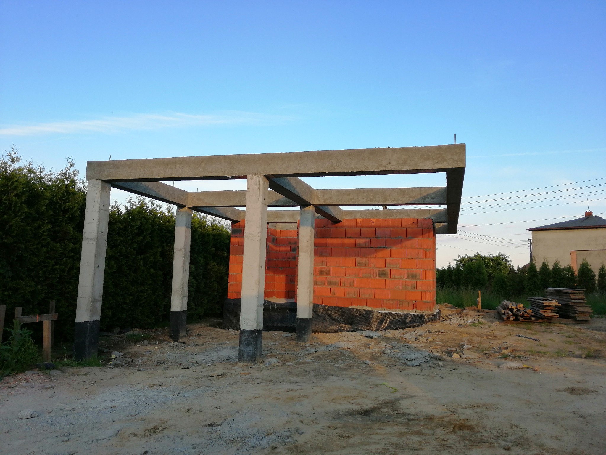 Wznoszenie ścian z czerwonej cegły na fundamencie z izolacją, widoczne betonowe słupy i elementy konstrukcyjne pod zadaszenie, teren budowy w trakcie prac.