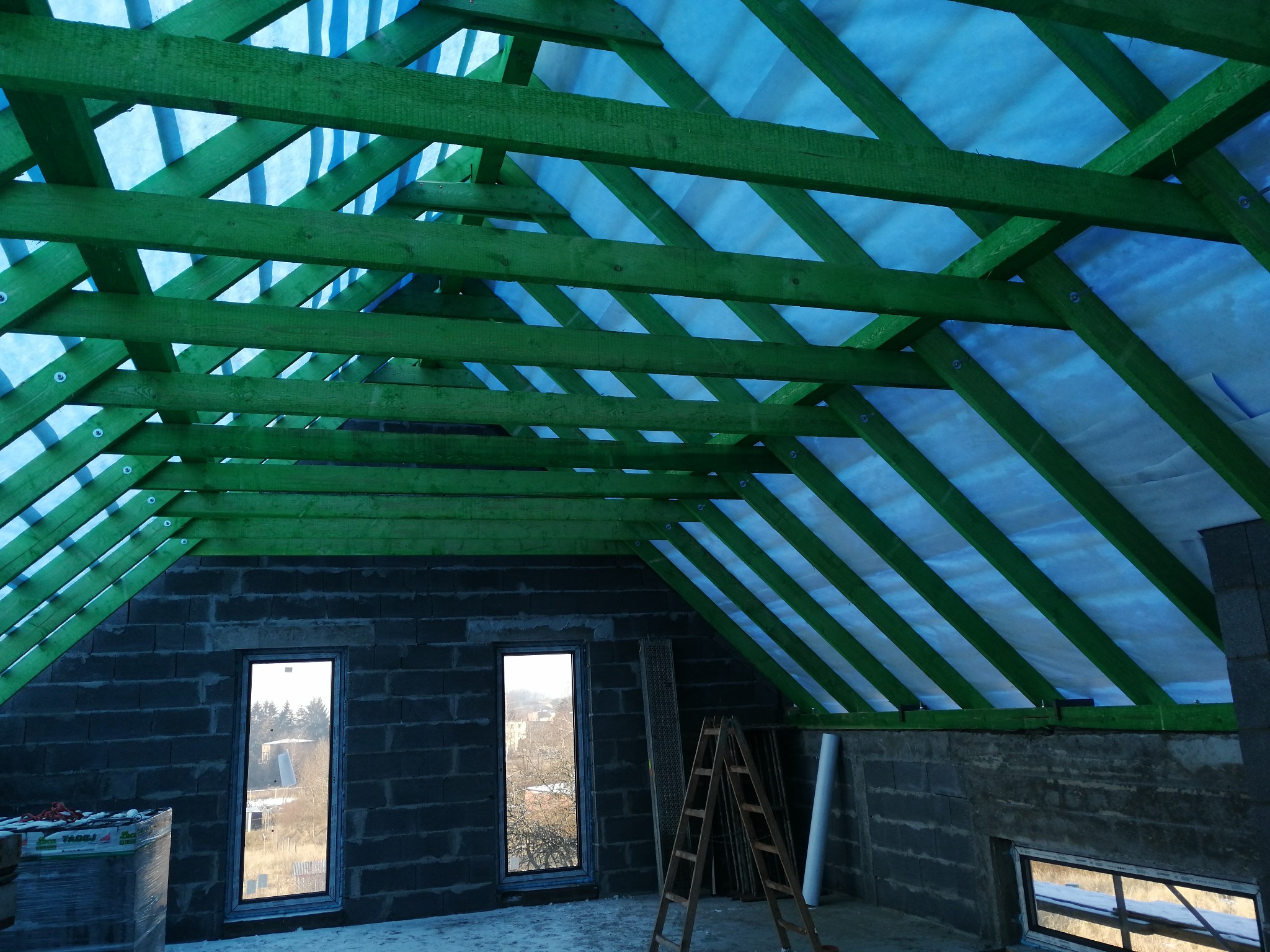 Wnętrze w trakcie budowy, widoczne ściany z bloczków, drewniana konstrukcja dachu pomalowana na zielono z niebieską membraną, okna wstawione, drabina oparta o ścianę.