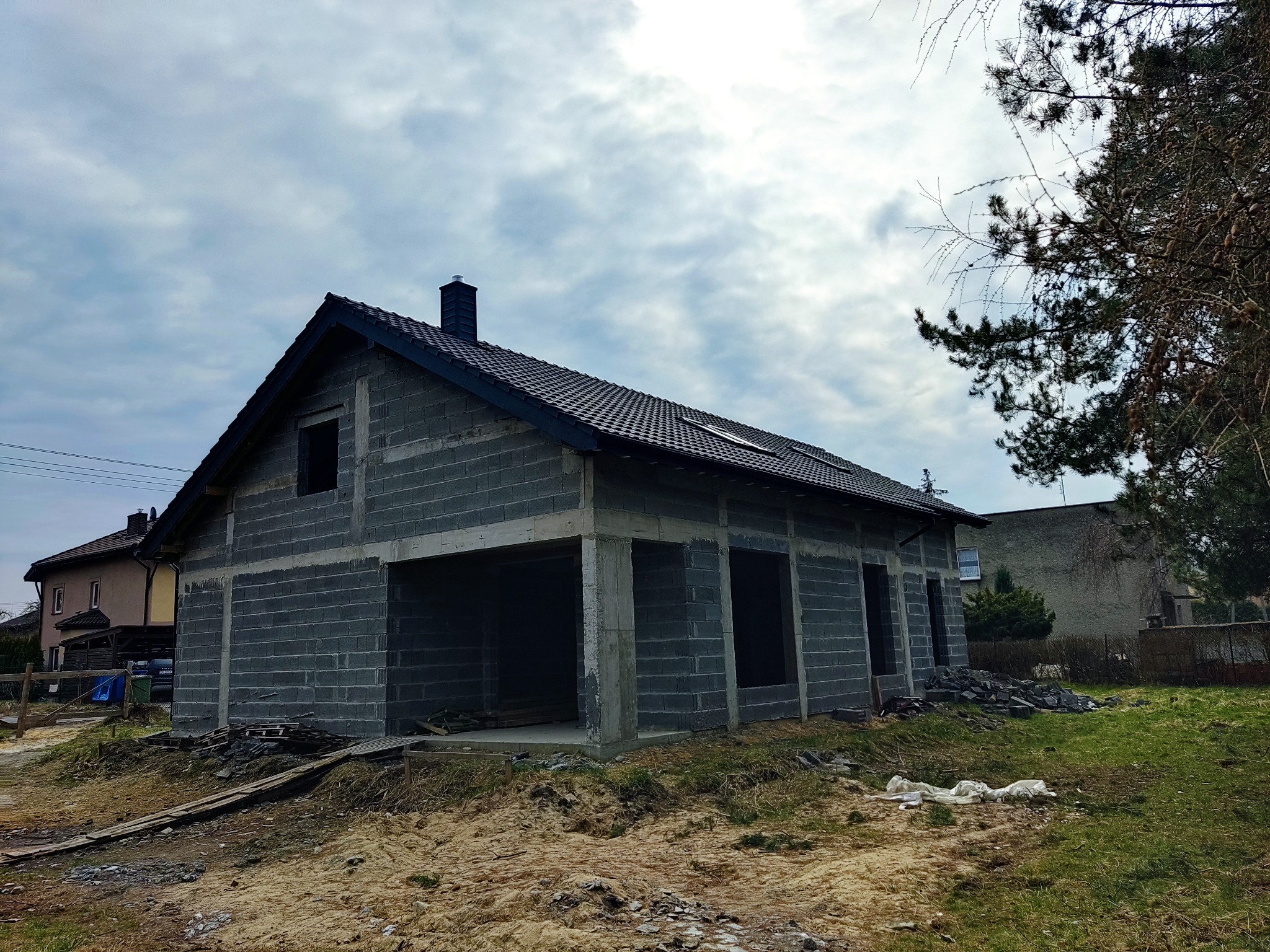 Surowy stan domu z bloczków betonowych z ciemną dachówką, widoczne otwory okienne i garażowe, otoczenie placu budowy z gruzem i drewnianymi deskami.
