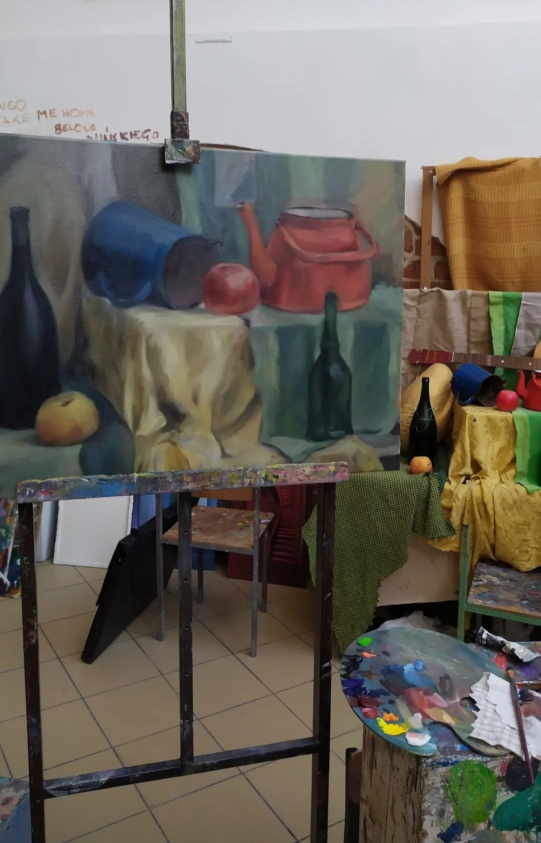 Martwa natura w trakcie tworzenia w Wołczkowie: niedokończony obraz na sztaludze przedstawia dzbanek, butelki i owoce, paleta malarska z farbami w pierwszym planie.