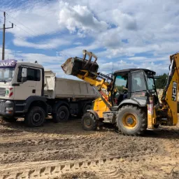 Żółta koparko-ładowarka JCB ładuje ziemię do wywrotki MAN na nierównym, piaszczystym terenie budowy pod błękitnym niebem z chmurami.