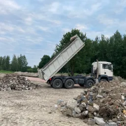 Ciężar&oacute;wka z podniesioną skrzynią wywrotki, wysypująca gruz na plac budowy, widoczne dwie pryzmy gruzu, w tle drzewa i pochmurne niebo.