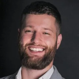 Uśmiechnięty mężczyzna z brodą w szarej marynarce na ciemnym tle, prezentujący się profesjonalnie.