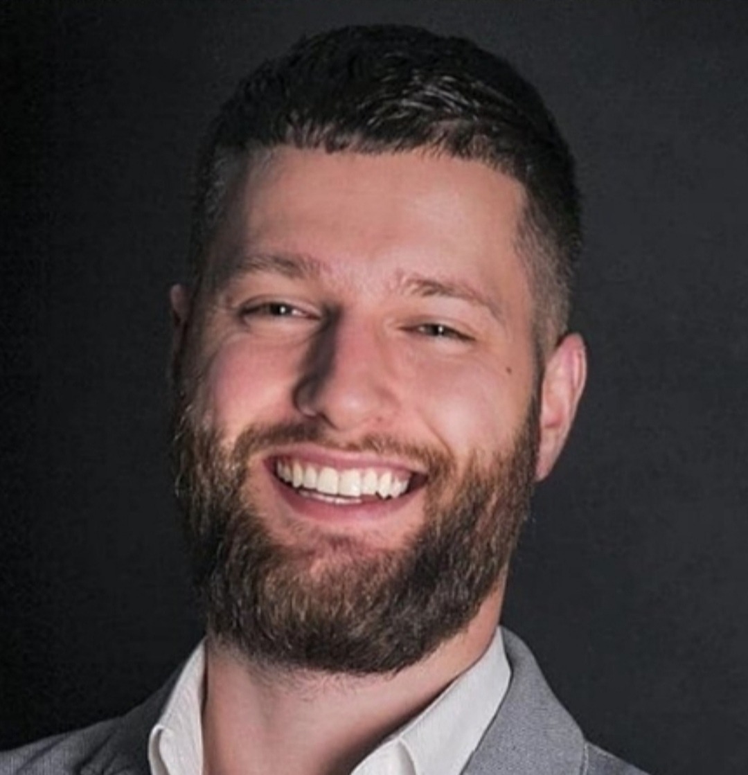 Uśmiechnięty mężczyzna z brodą w szarej marynarce na ciemnym tle, prezentujący się profesjonalnie.