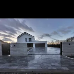 Nowoczesny dom jednorodzinny z podjazdem wyłożonym szarą kostką brukową, widok z poziomu gruntu na otwartą bramę wjazdową i minimalistyczną architekturę budynku w tle, pochmurne niebo.