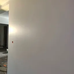 Świeżo wyszpachlowana ściana pomalowana na jasny odcień szarości, widoczne gniazdka elektryczne zabezpieczone czerwonymi zaślepkami, podłoga zabezpieczona folią malarską.