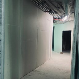 Budland - Wnętrze w trakcie prac wykończeniowych: zamontowane płyty gipsowo-kartonowe na ścianach, widoczne metalowe profile konstrukcyjne, instalacje pod sufitem i surowy beton na podłodze, korytarz...