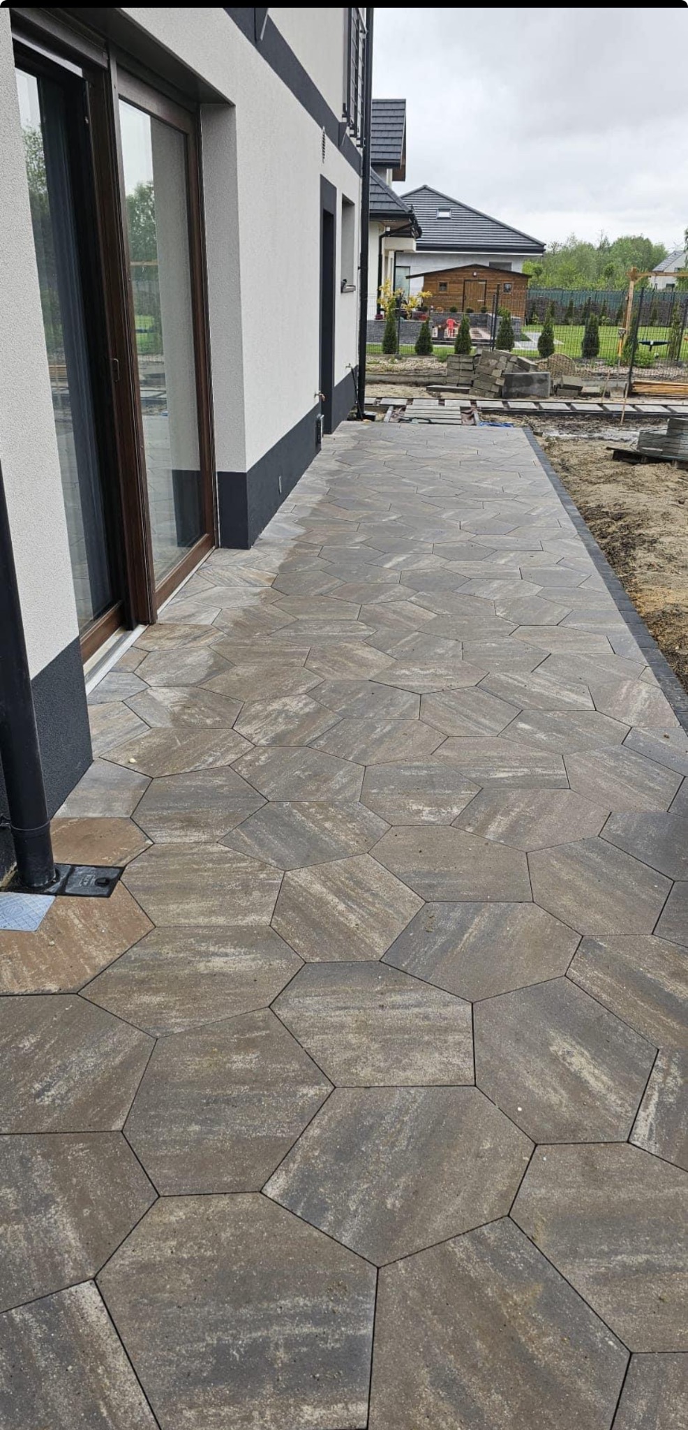 Ułożona ścieżka z szarej kostki brukowej o heksagonalnym kształcie, prowadząca wzdłuż ściany budynku z oknem przesuwnym, widoczne pozostałości po pracach brukarskich.