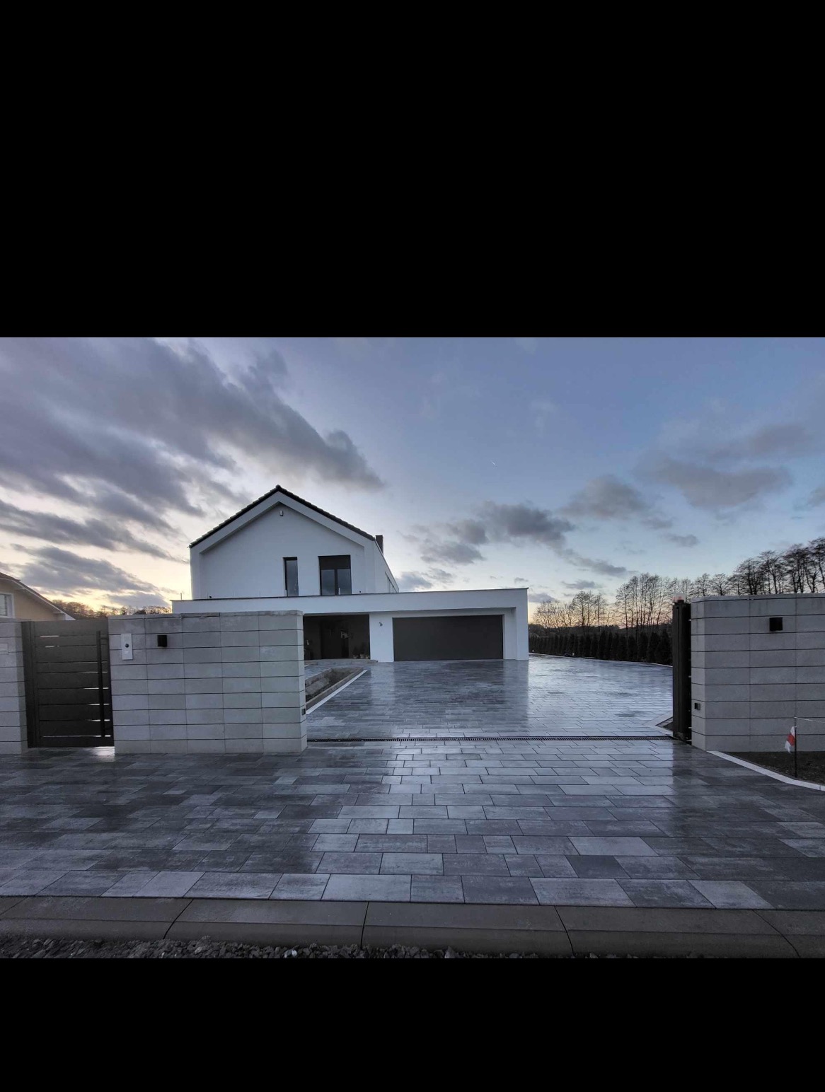Nowoczesny dom jednorodzinny z podjazdem wyłożonym szarą kostką brukową, widok z poziomu gruntu na otwartą bramę wjazdową i minimalistyczną architekturę budynku w tle, pochmurne niebo.