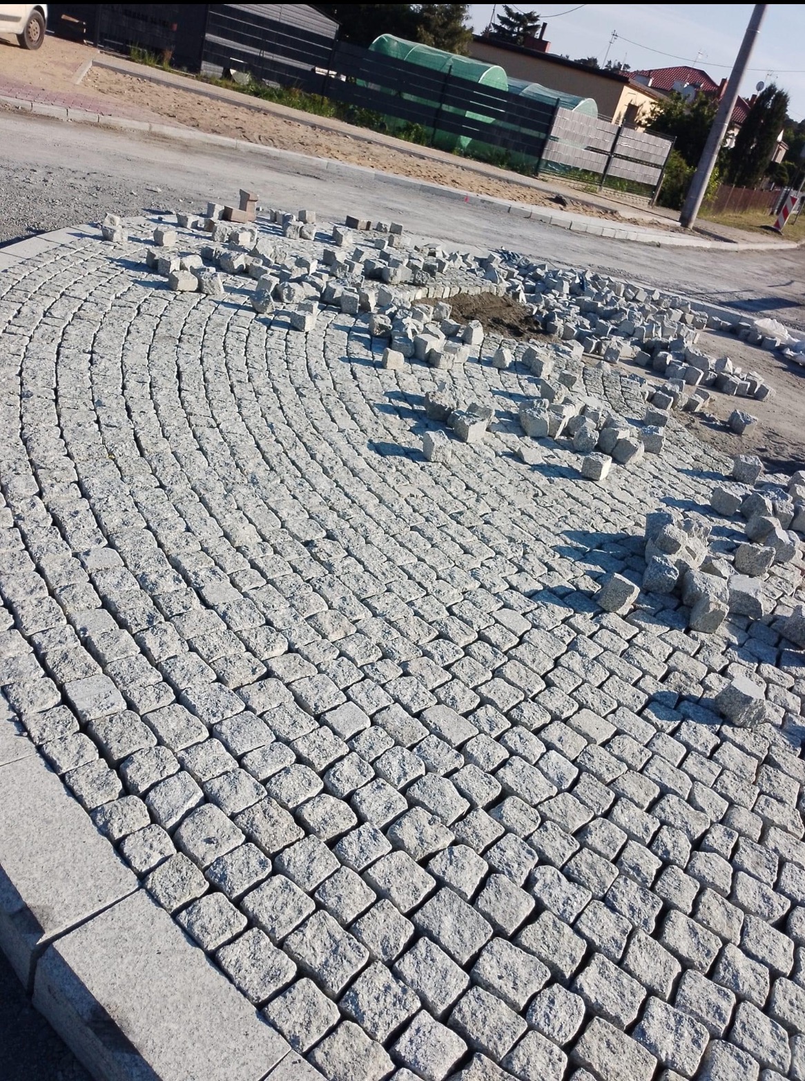 Fragment ulicy z ułożoną szarą kostką brukową w kształcie łuku, z widocznymi jeszcze nieułożonymi kostkami i niewielką ilością piasku, w tle droga i zabudowania.